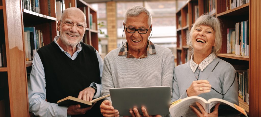 Two older adult men and one woman are smiling together in a library